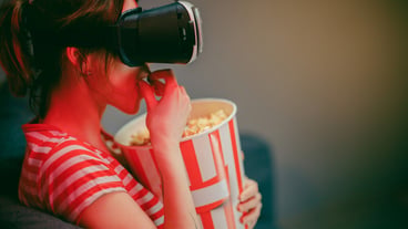 Woman wear VR headset and watch movie with popcorn at night. American woman sitting on the sofa in the VR glasses and watching something while eating popcorn. Indoor