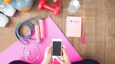 Young woman with cell phone sitting near weights on yoga mat, top view.