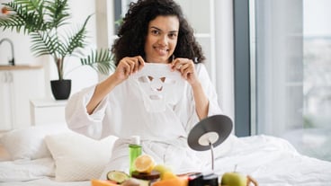 Woman holding mask over fruits and cosmetics on tray.