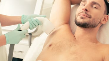 Close up photo of a young calm man lying with his eyes closed and smiling when professional doctor removing hair on his armpit