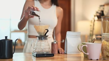 Closeup of hand pouring coffee grounds into a dripper on a wooden kitchen counter