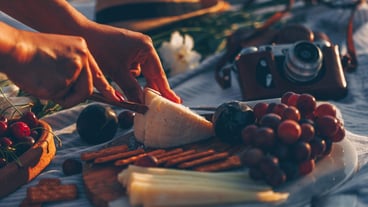 woman slicing cheese on wooden cutting board with cheese and fruits on it and camera, hat and flowers in beach.