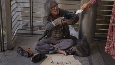 Homeless man is sitting down on walkway in town. He is holding hat and receive dollar.