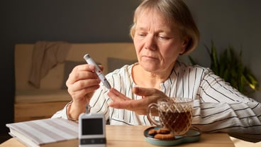 A senior woman is measuring her blood sugar levels with a glucometer at a wooden table Next to her a cup of tea and a plate of cookies sit creating a warm atmosphere