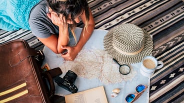 High angle view of young woman lying on carpet with coffee cup and looking at atlas. Young woman planning for location on map with magnifying glass on paper. Woman planning her vacation looking at map