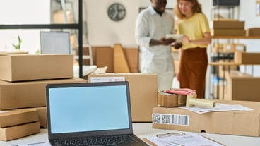 Laptop with white empty screen standing on desk among packed boxes and documents against two young intercultural managers using tablet