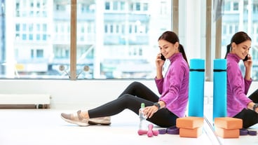 Side view of a young beautiful fitness woman in sportswear sitting on the floor in fitness studio or gym and talking on mobile phone, resting after workout. Sport, wellness and healthy lifestyle