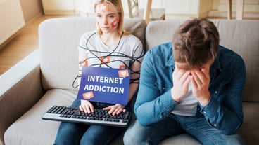 The young woman is addicted to social media. She sits numbly on the couch. Her body is entangled in a cable. Her hands are on the keyboard. A desperate young man sits on the couch and cries.