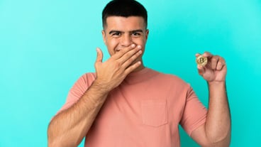 Handsome young man holding a Bitcoin against an isolated blue wall, happy and smiling, covering his mouth with his hand