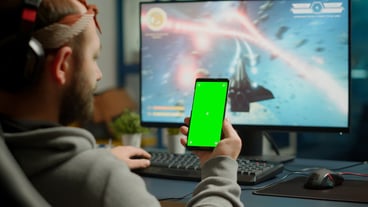 Focus on the gamer holding the phone with the green screen mock-up chroma key display while playing games on a powerful computer streaming online competition. Gamer using an isolated desktop to...