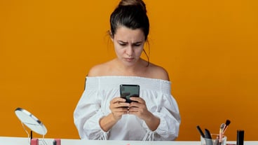 Annoyed beautiful girl sits at table with makeup tools holding and looking at phone isolated on orange wall