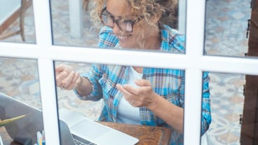 Middleaged woman with curly hair and glasses seen through a window while using a laptop for a video call smiling and talking to colleagues during an online business meeting in her home office
