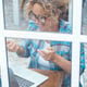 Middleaged woman with curly hair and glasses seen through a window while using a laptop for a video call smiling and talking to colleagues during an online business meeting in her home office