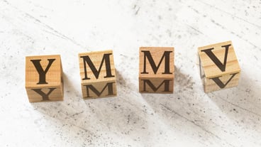 Four wooden cubes with letters YMMV meaning Your Mileage May Vary on white working board..