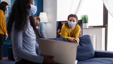 Happy cheerful woman holding a bottle of beer looking at a laptop talking to an African friend in the living room of the apartment with a mask to prevent the spread of coronavirus in times of a global pandemic....