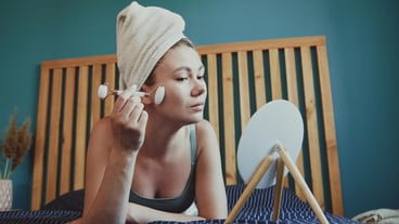 Young woman using jade facial roller for face massage lying on bed in bedroom looking in the mirror