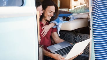 Happy couple using laptop computer inside minivan during road trip vacation - Focus on man face