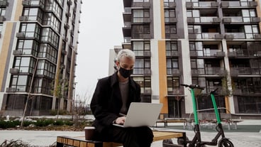 Concentrated freelancer man in black face mask working outside in park and using his modern laptop. He sits on bench and writes a program. Two electric scooters near the bench. Apartment blocks.