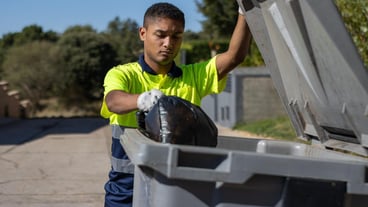 A municipal worker throwing a bag of garbage into a large dumpster