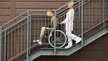 Side view of smiling friendly female rehabilitation centre worker pushing wheelchair with patient wh