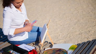 Female painter holding a box with paints while sitting on a wooden chaise lounge on the beach with canvas and palette on it
