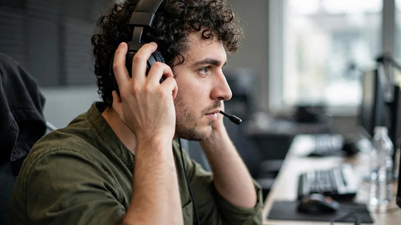 A young man with curly hair wearing a headset with a microphone in front of a computer setup, looking toward the screen.