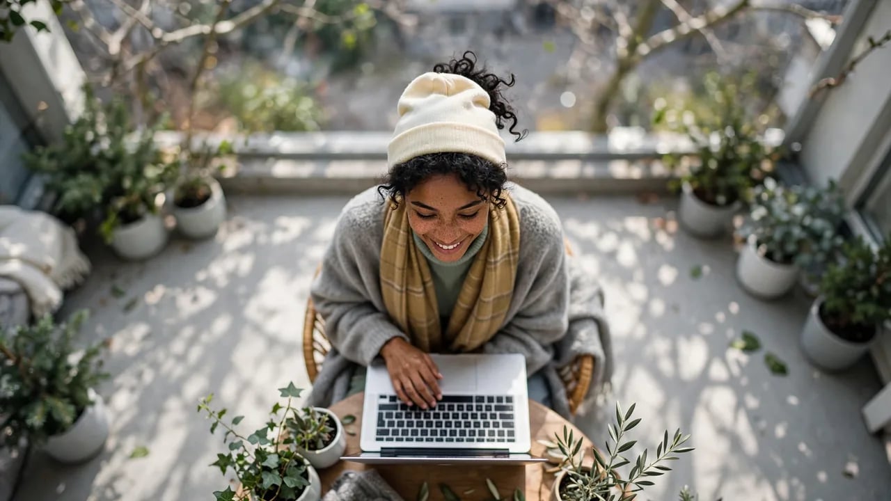 A woman wearing a beanie and scarf smiling while typing on a laptop on a balcony surrounded by potted plants.