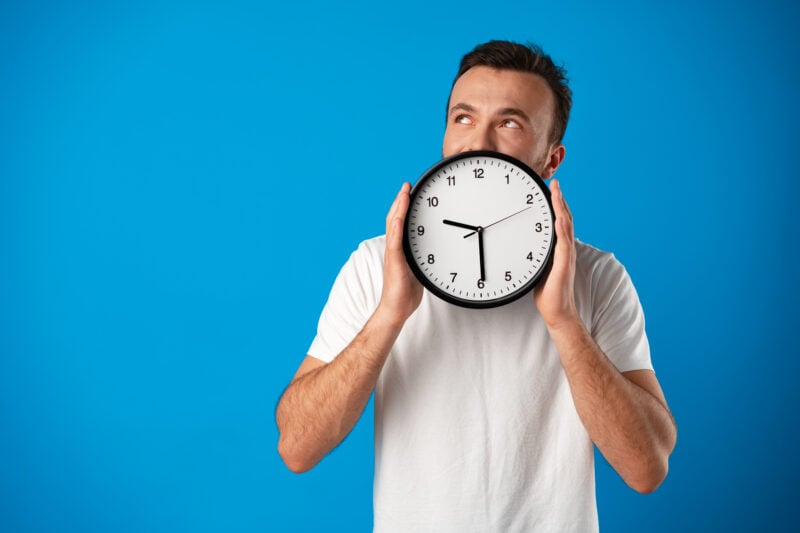 Handsome young man in white t-shirt posing with clock against blue background
