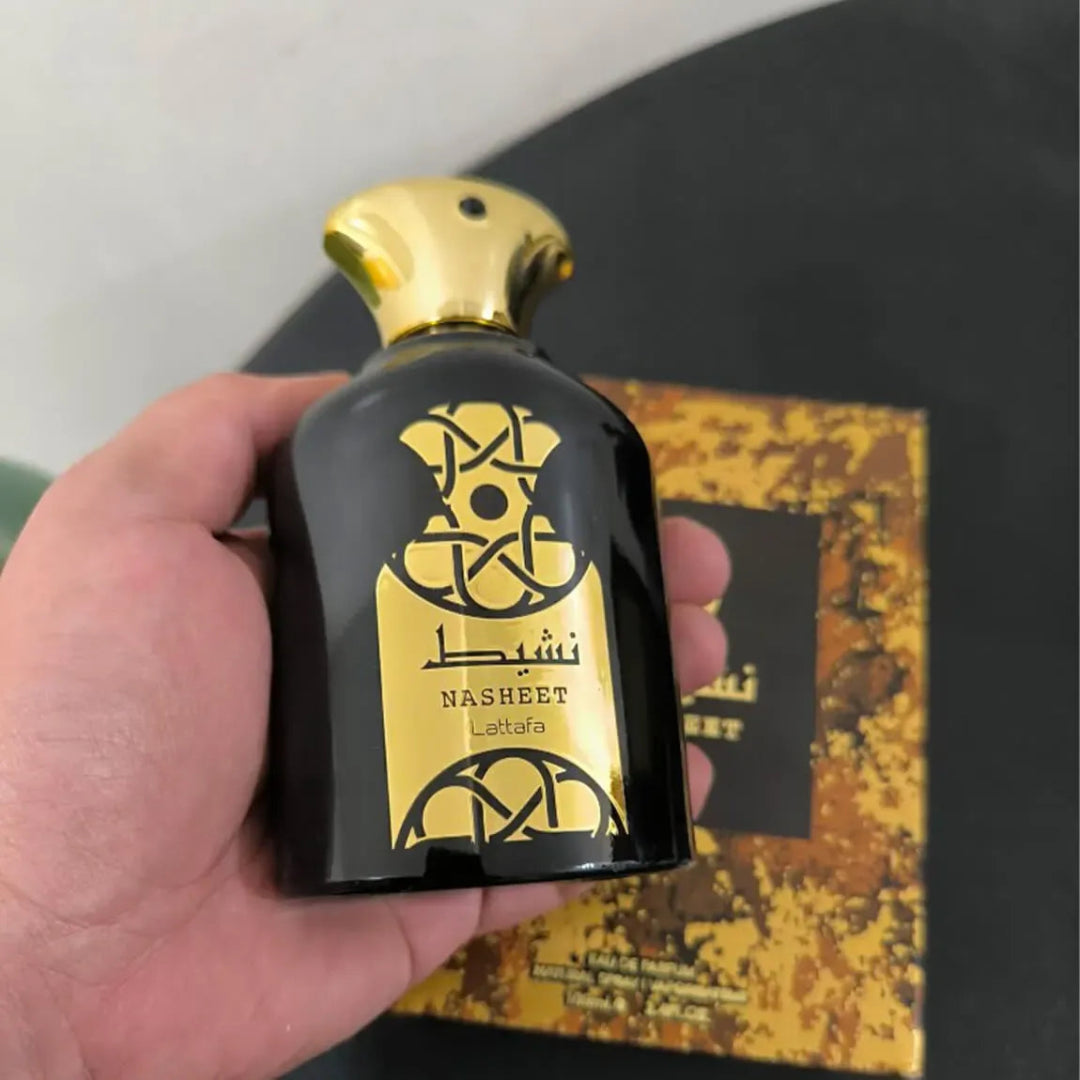 Hand holding a black and gold perfume bottle labeled 'Nasheet Latafa' with a blurred background.