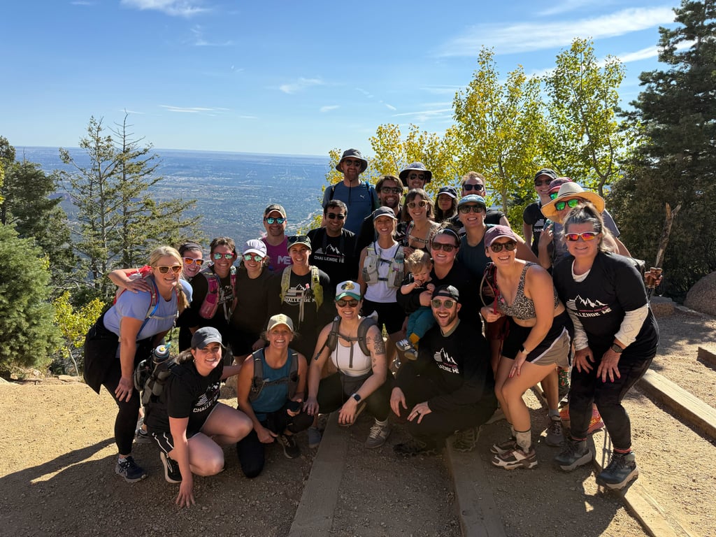 A large group of smiling hikers poses together at the top of a scenic mountain trail overlooking a vast valley on a sunny day.