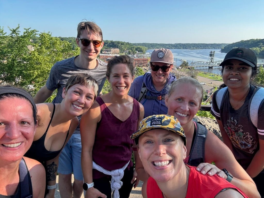 A group of smiling, active friends poses for a selfie outdoors on a sunny day with a river and lift bridge in the background.