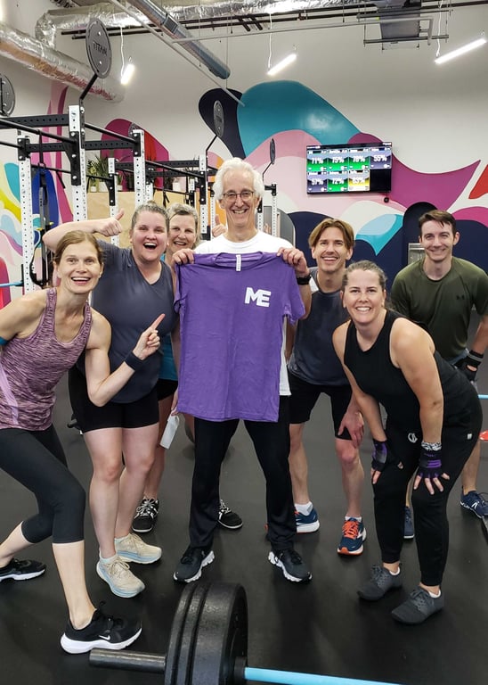 A smiling group of fitness members poses together in a gym, celebrating as an older man holds up a purple t-shirt with a white "ME" logo.