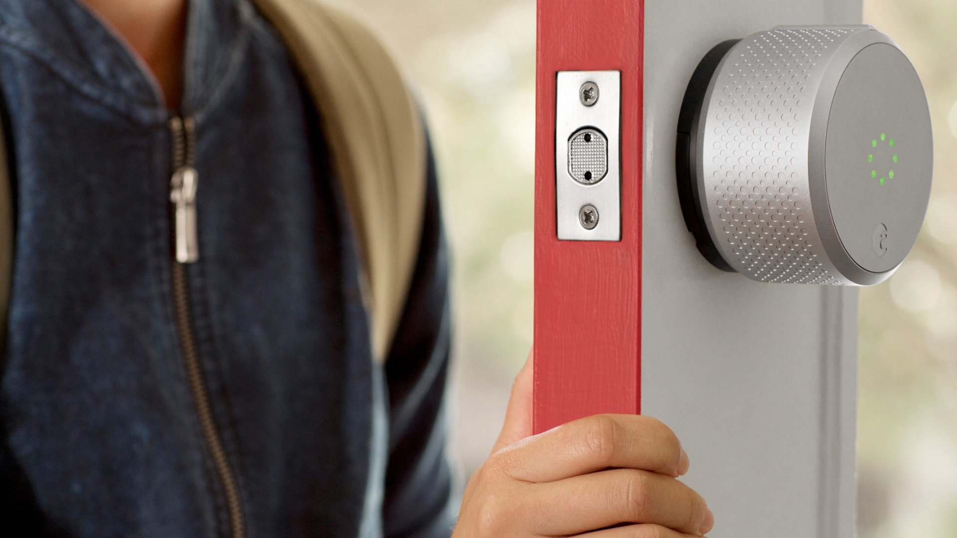 Close-up of the silver August Smart Lock mounted on a red door frame, glowing with a green status ring as a person in a denim