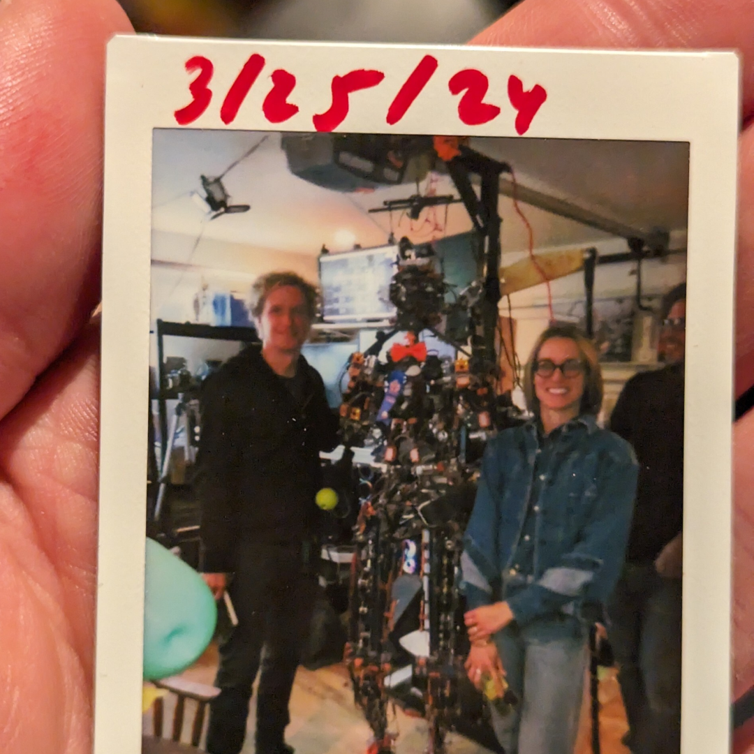 Polaroid photo dated 3/25/24 of the Kind Humanoid team posing with their early bipedal robot prototype in a workshop