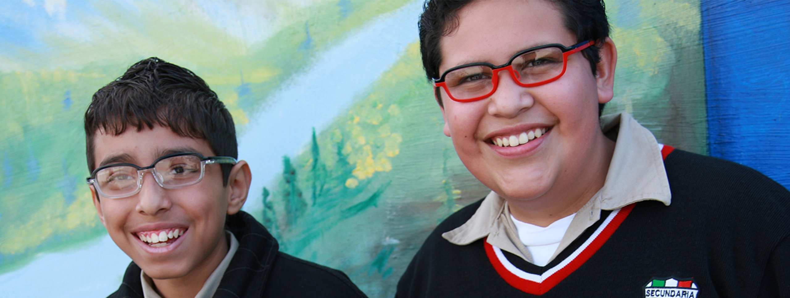 Two smiling Mexican students wearing Verbien Augen Optics affordable prescription eyewear in clear and red frames in front of