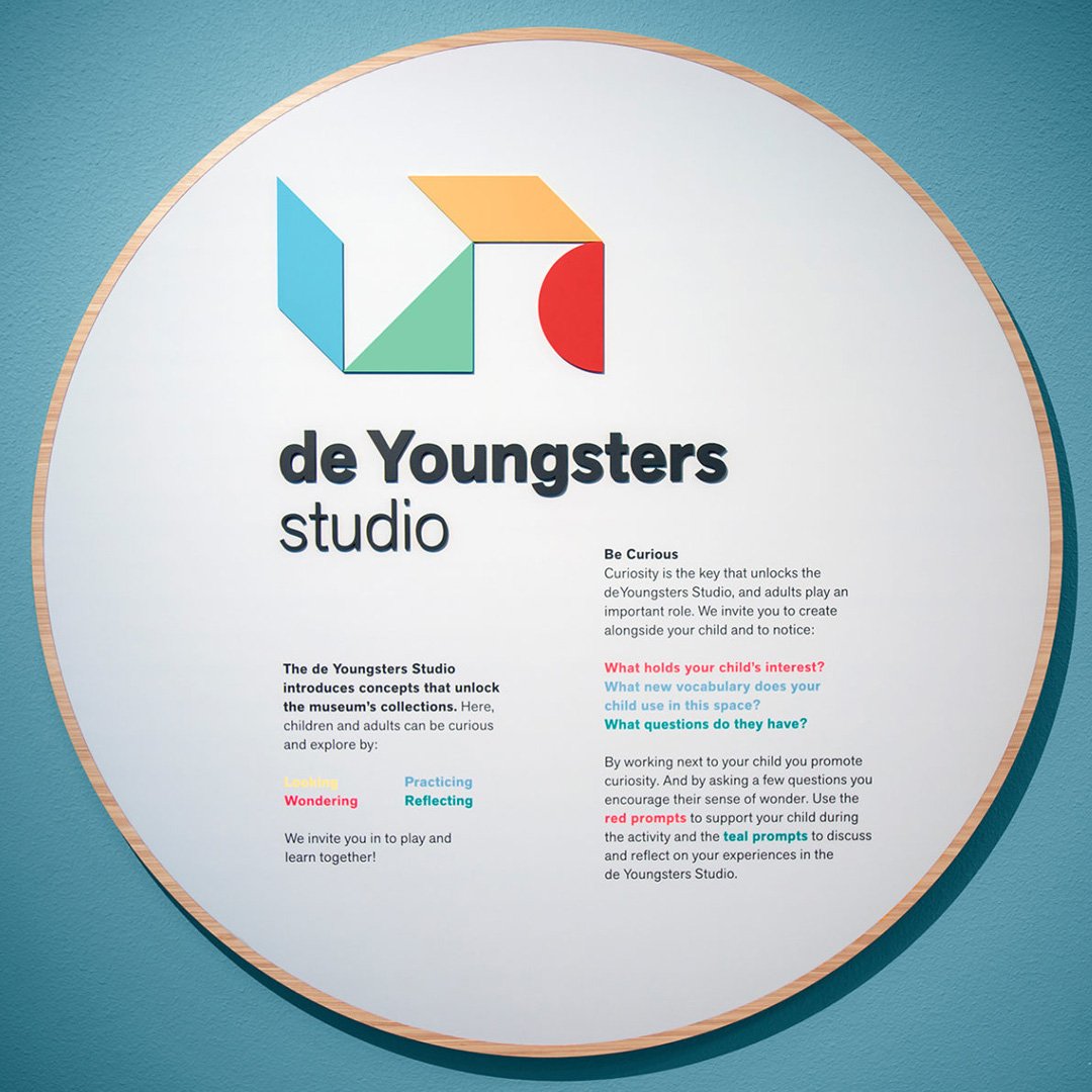 Round wall-mounted de Youngsters studio signage with colorful geometric logo and Be Curious introduction text on a teal museu