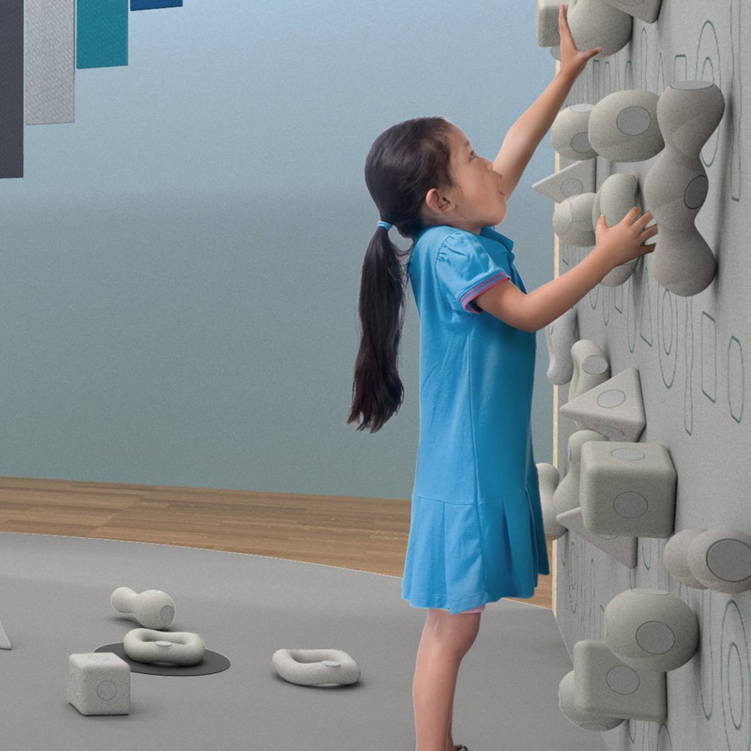 Young girl in blue dress reaches up to climb a soft sculptural rock-shaped climbing wall in a De Youngsters Studio playroom w