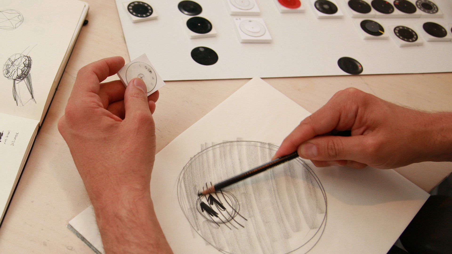 Designer Yves Behar sketching VUE watch dial concepts in pencil, holding prototype model with watch face mockups arrayed on w