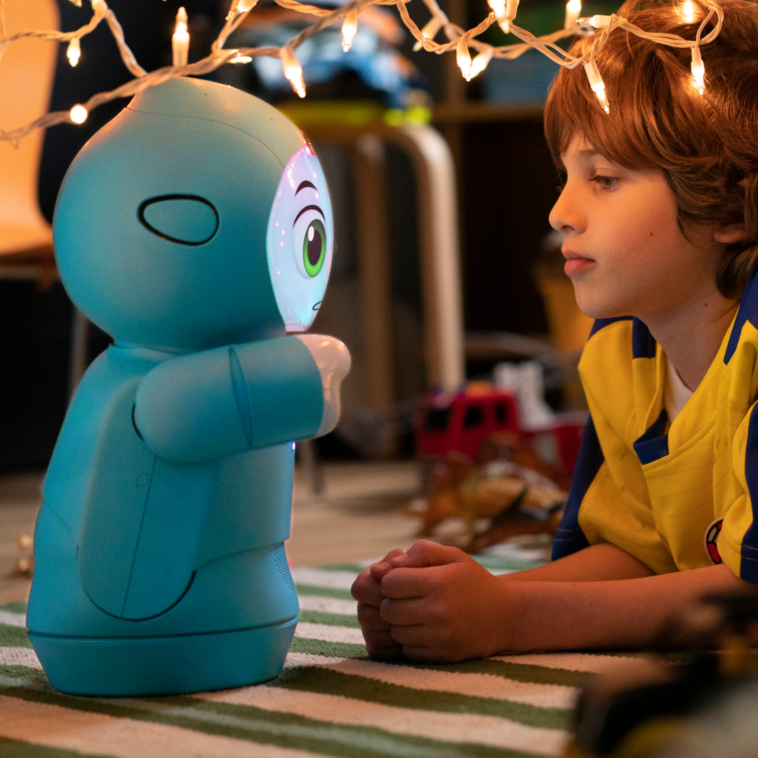 Boy sitting next to blue robot with friendly face in indoor setting