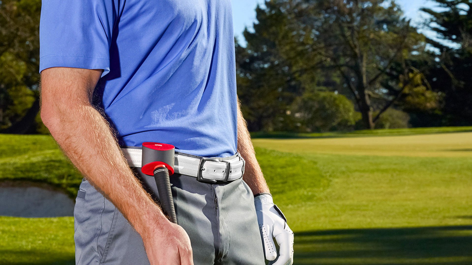 Golfer in blue shirt and grey pants on the fairway wearing the Game Golf red and silver swing-tracking sensor clipped to his