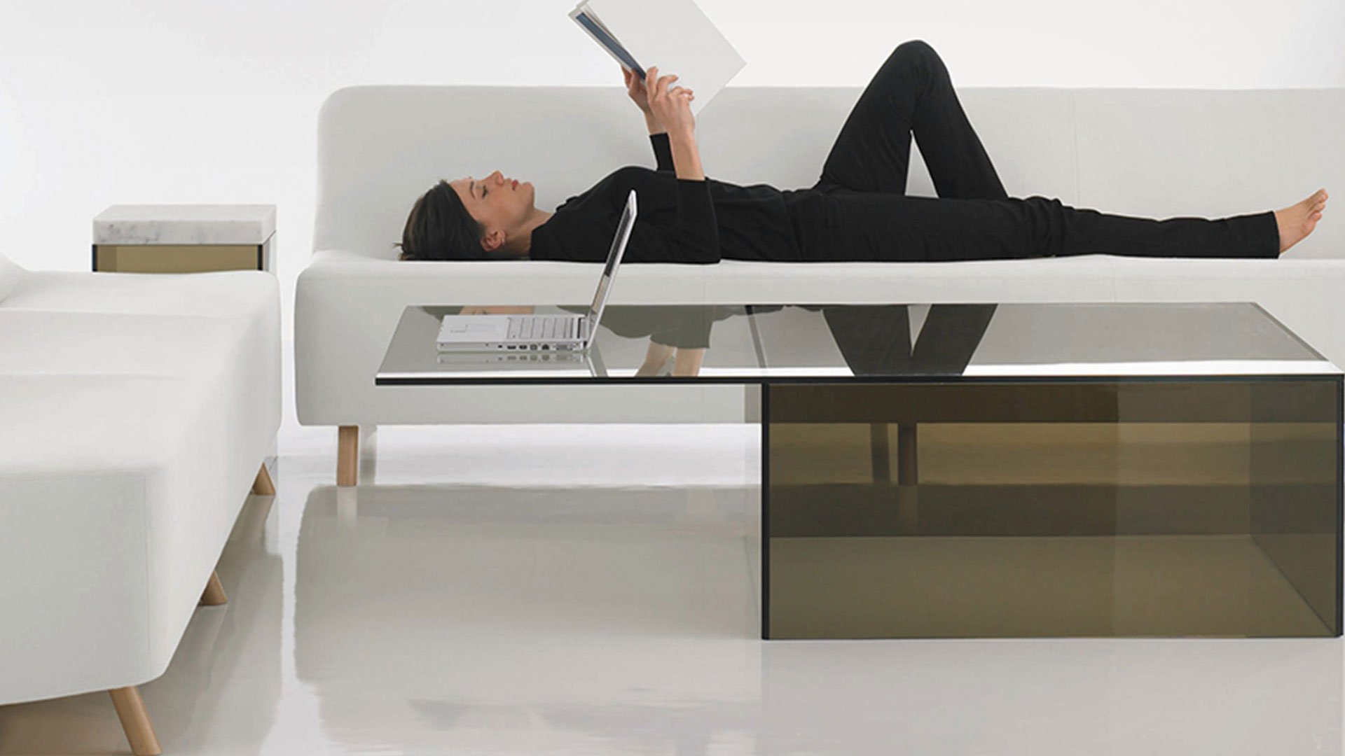 HBF modern living room featuring woman in glasses reading on a white sofa beside a glass coffee table