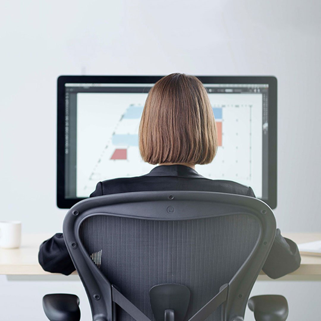 Woman with bob haircut seated in a Herman Miller Aeron mesh-back office chair working at a large desktop monitor