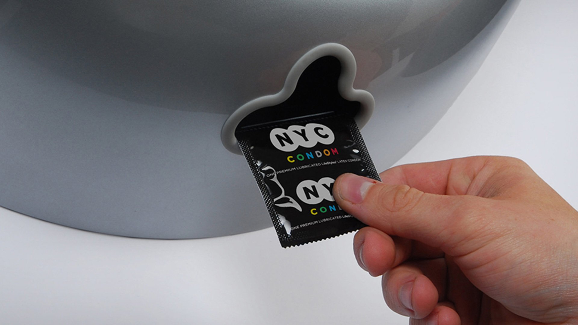 NYC Condom packet held in hand showing colorful design against a gray background with toilet lid