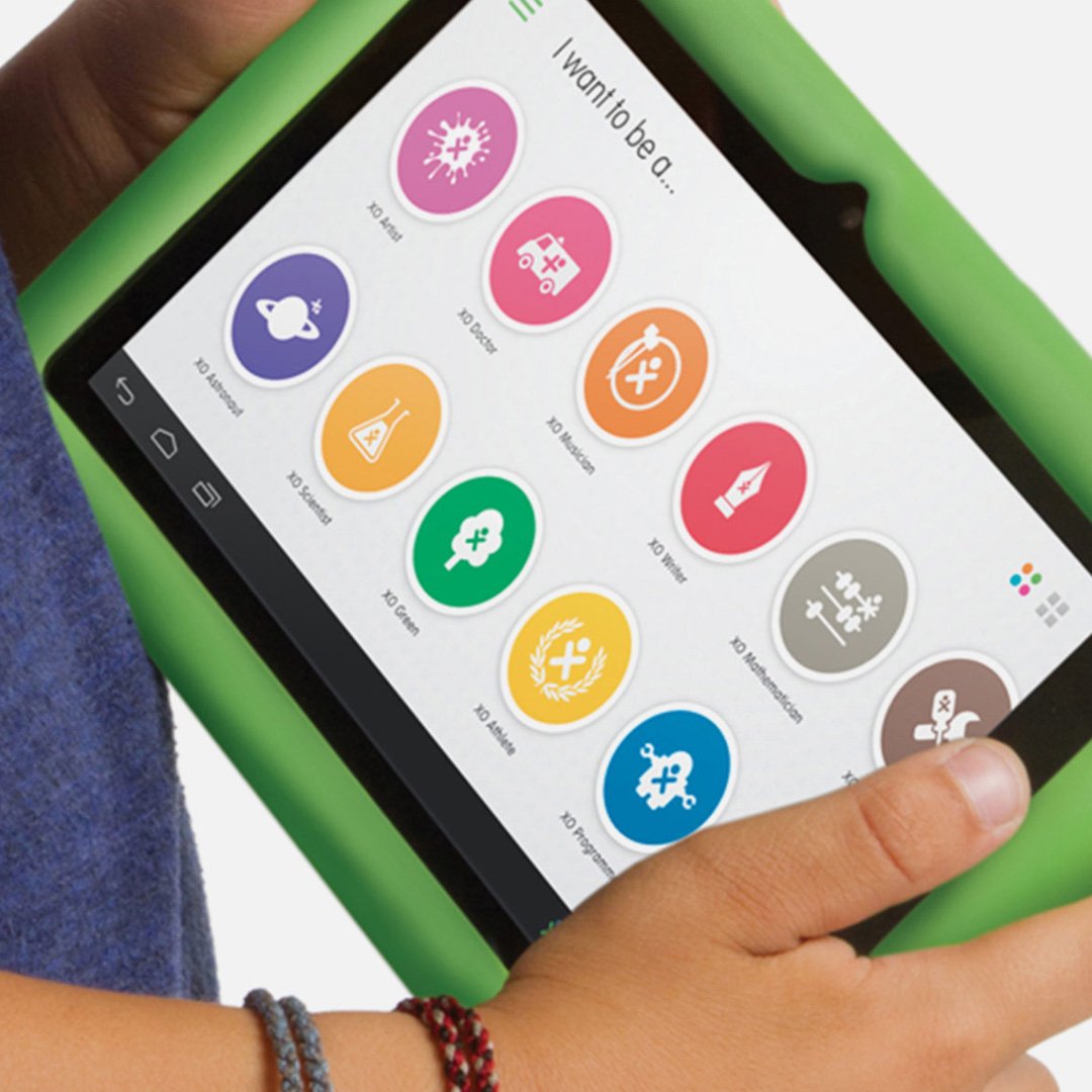 Child holding OLPC XO green tablet showing colorful "I want to be a..." activity icons including artist, doctor, musician, wr