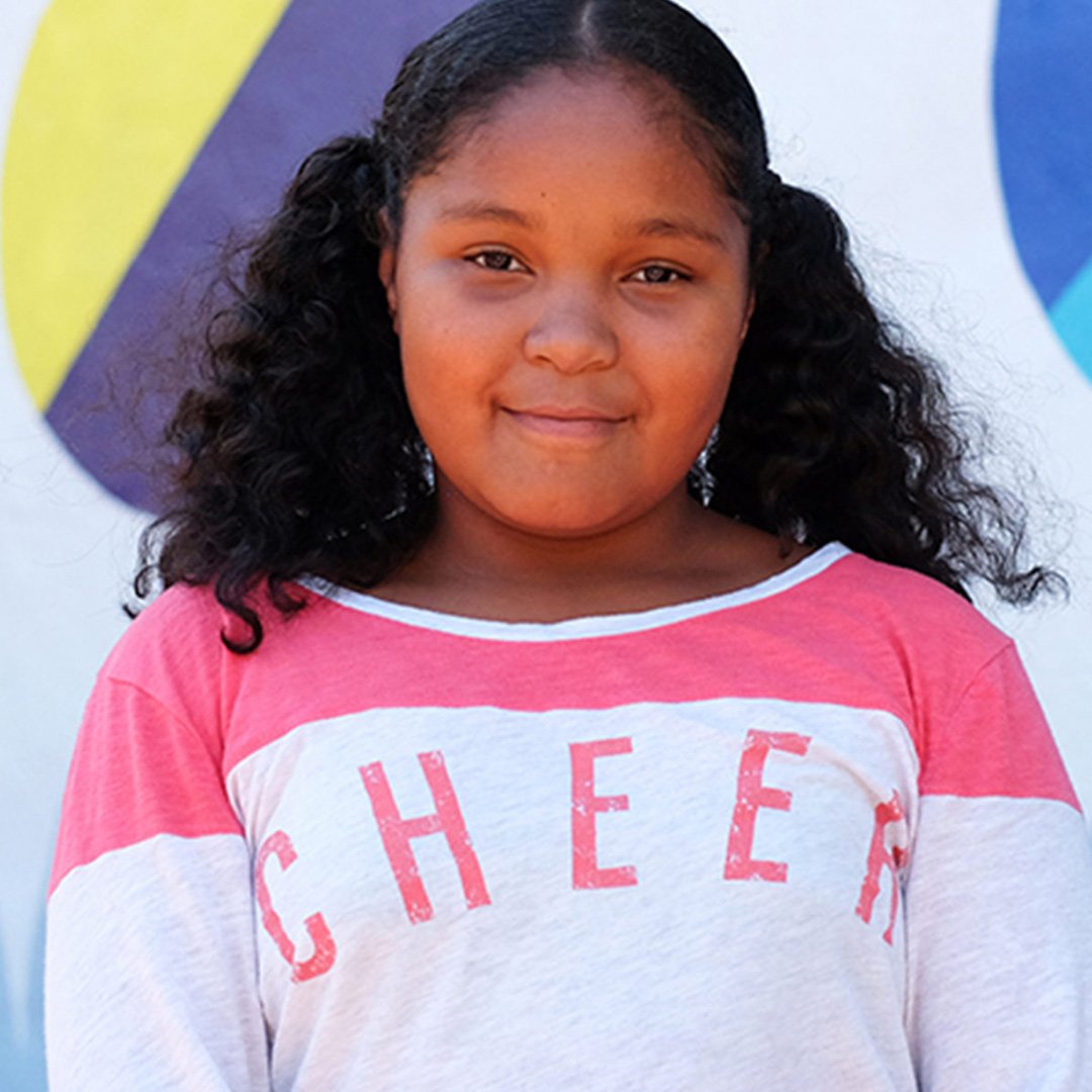 Smiling young Boys & Girls Club member wearing a pink and white CHEER shirt in front of the colorful Willie Mays mural backdr