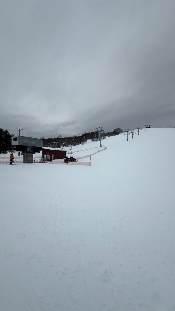 A snowy ski slope with a chairlift running up the hill, surrounded by a cloudy, overcast winter landscape.