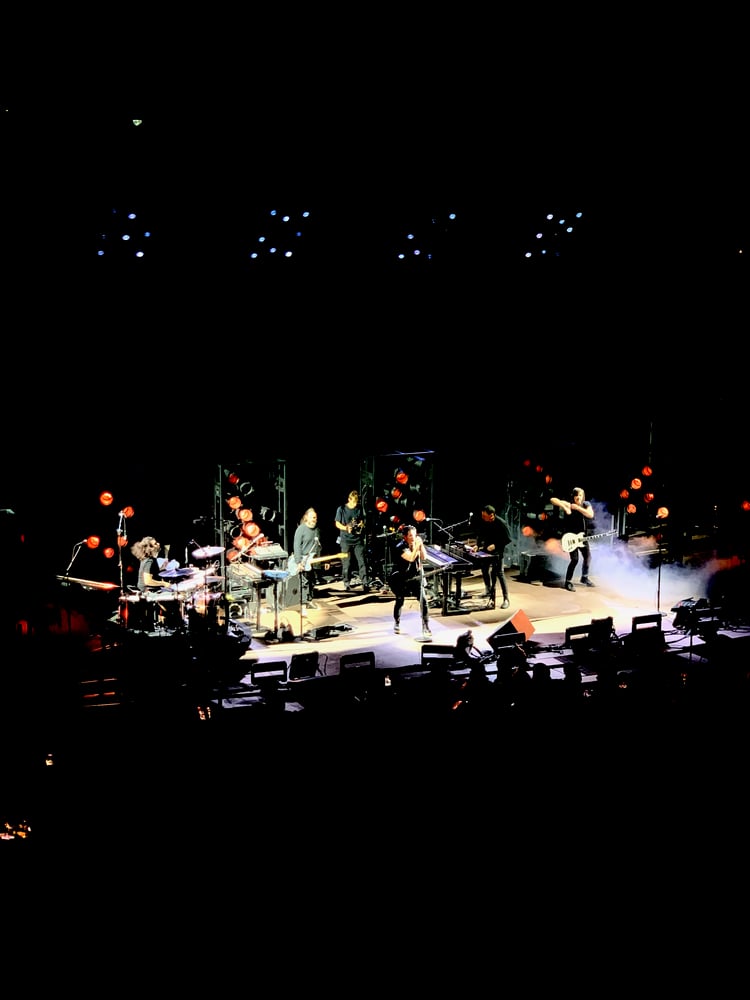 The image shows a live music performance on a stage with multiple musicians playing various instruments, including drums, keyboards, and guitars. The stage is dimly lit, with colorful lights illuminating the performers. The audience can be seen in the foreground, creating an energetic and immersive atmosphere.
