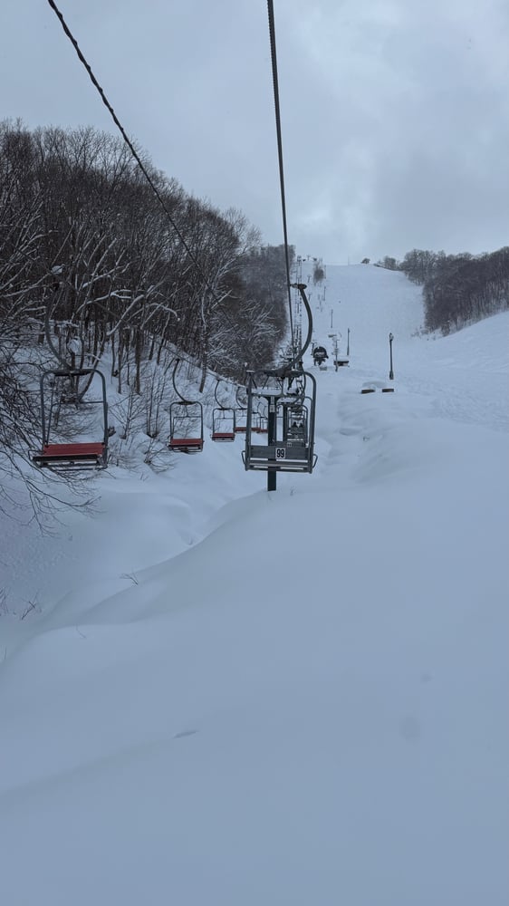 A snow-covered ski resort with a chairlift carrying skiers up the slope, surrounded by bare trees and a wintry landscape.