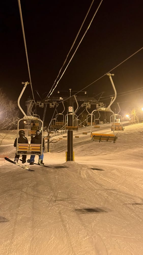 A night image of a ski lift and ski slope, with skiers riding the lift and snow-covered terrain below.