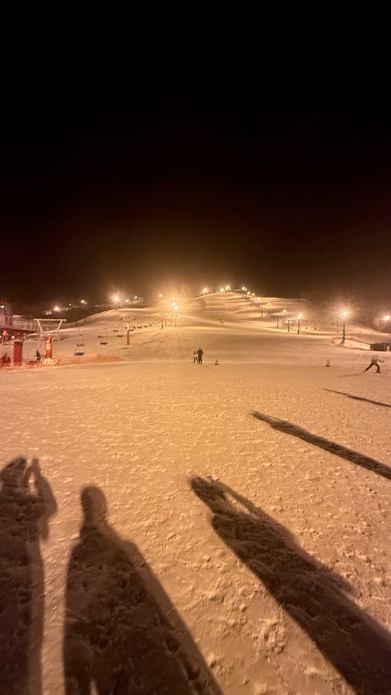 A snowy ski resort at night, with people skiing and snowboarding on the illuminated slopes. The image shows a panoramic view of the resort with a warm, golden glow from the lighting.